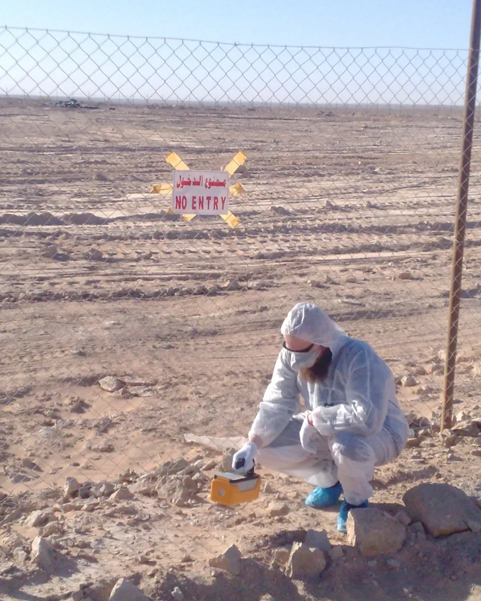 Technician in protective coveralls taking a ground sample by a fenced site